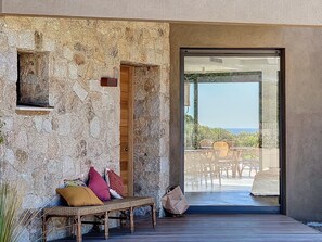 A rustic stone wall with a wooden bench and cushions, a door leading to an outdoor seating area with a view of the sea. - Superbe Villa vue Mer, Piscine Chauffée, Terrain de Pétanque (Corbara)
