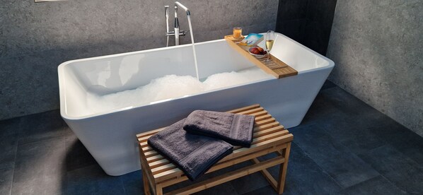 Et moderne badeværelse med et stort badekar, en træskammel og en bakke med en bog, frugt og et glas vin.