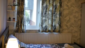 Ein Schlafzimmer mit Fenster, Heizkörper, Bett mit gemustertem Überwurf, Nachttischlampe, Regal mit Pflanzen und Vorhängen mit Blattmuster.