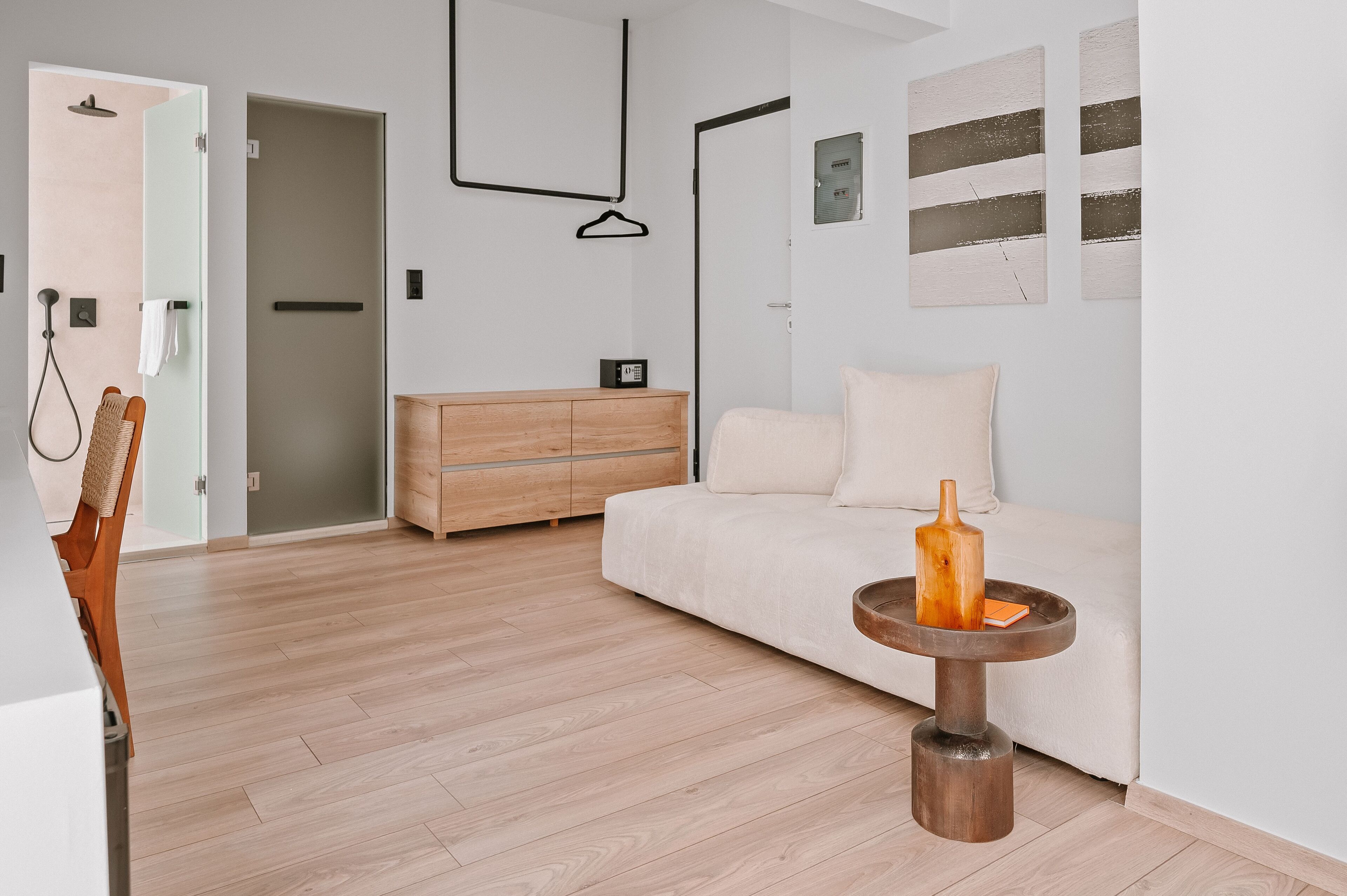 Habitación moderna y minimalista con piso de madera, sofá blanco, cómoda de madera y una pequeña mesa redonda con una botella y un libro.