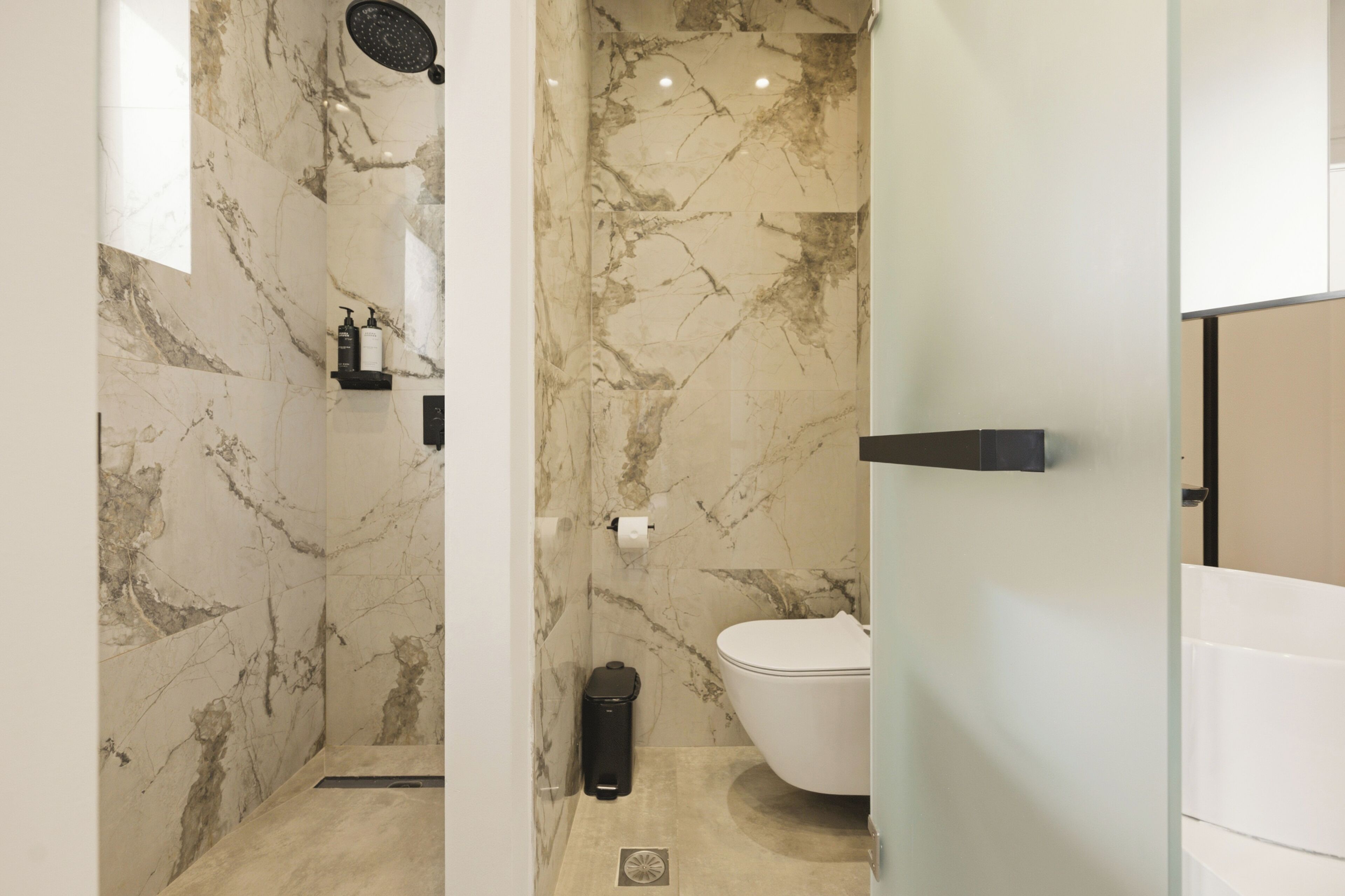 A bathroom with marble walls, a white toilet, a black trash bin, and a shower head.
