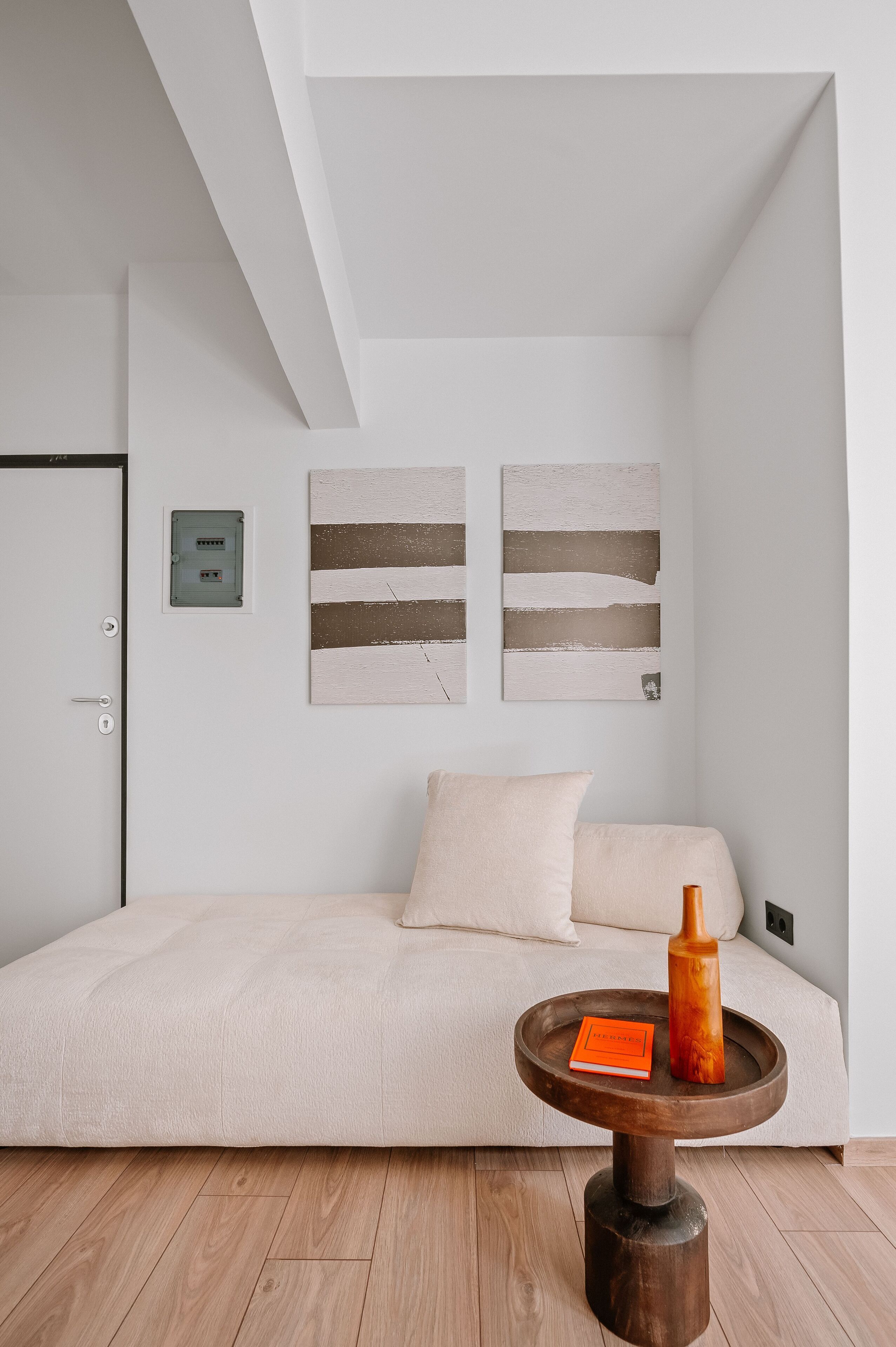 Une chambre à coucher au style minimaliste, avec un lit, une petite table en bois sur laquelle repose un livre, et des œuvres d’art abstraites accrochées au mur.