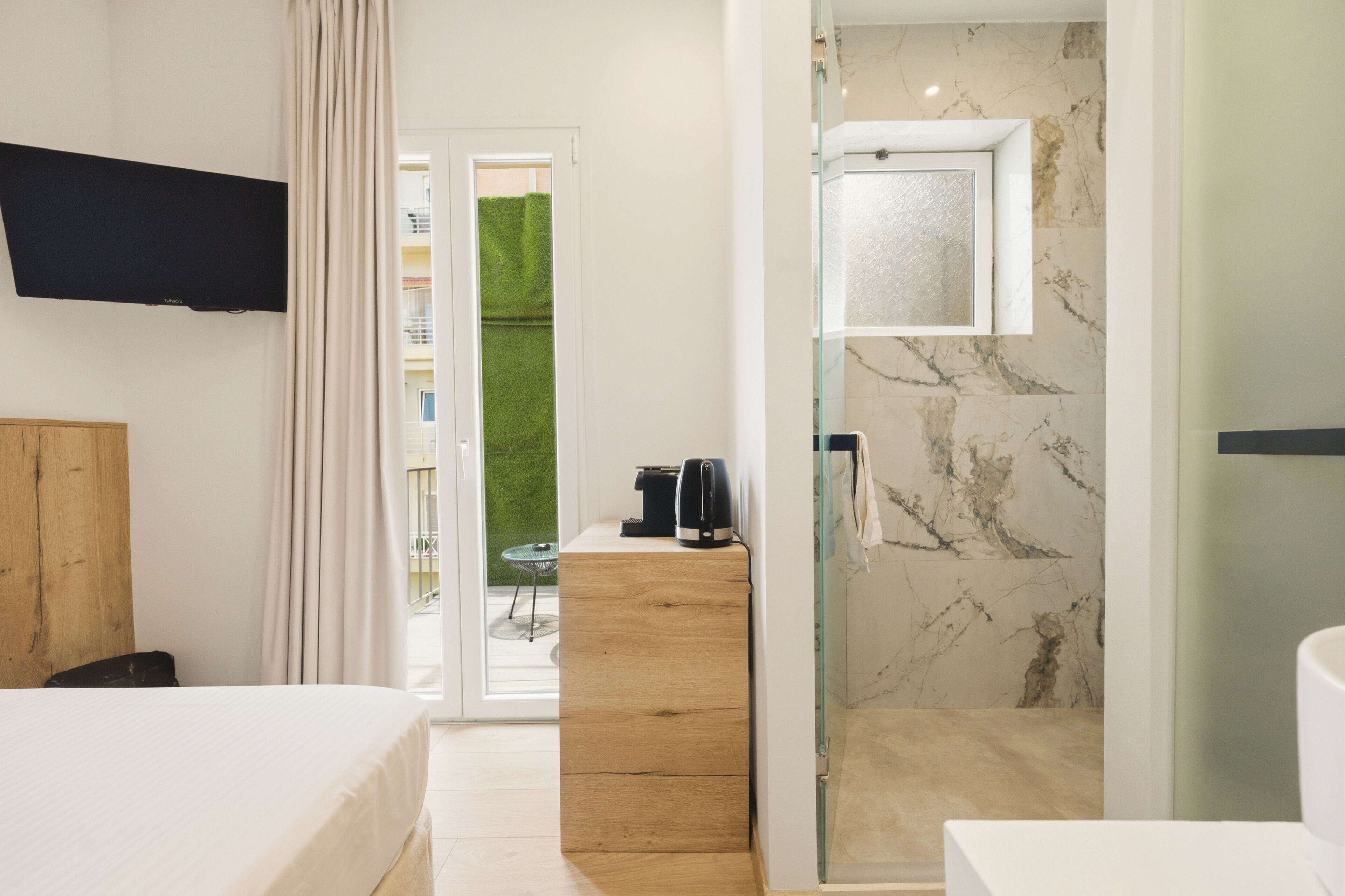 Habitación de hotel con cama, cómoda de madera, cafetera, televisor, cabina de ducha de vidrio y vista a un césped verde.