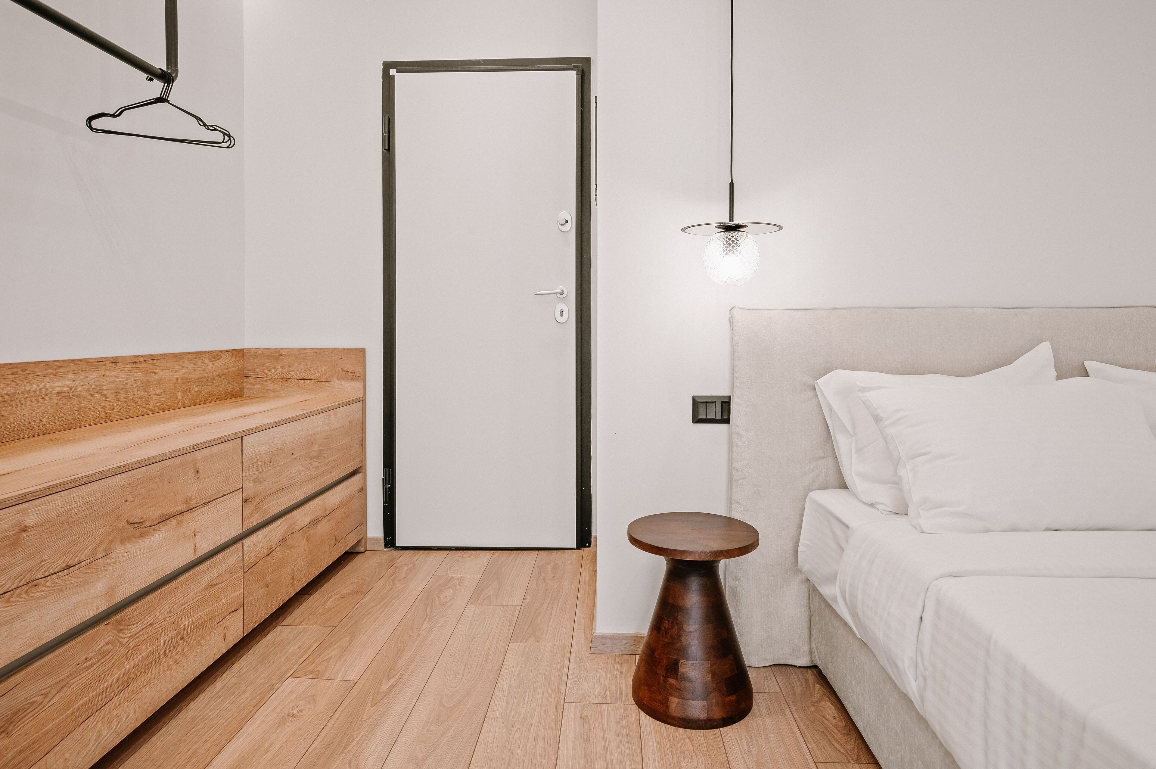 Un dormitorio moderno con un banco de madera, una mesa redonda de madera, una cama con ropa blanca y una lámpara colgante.