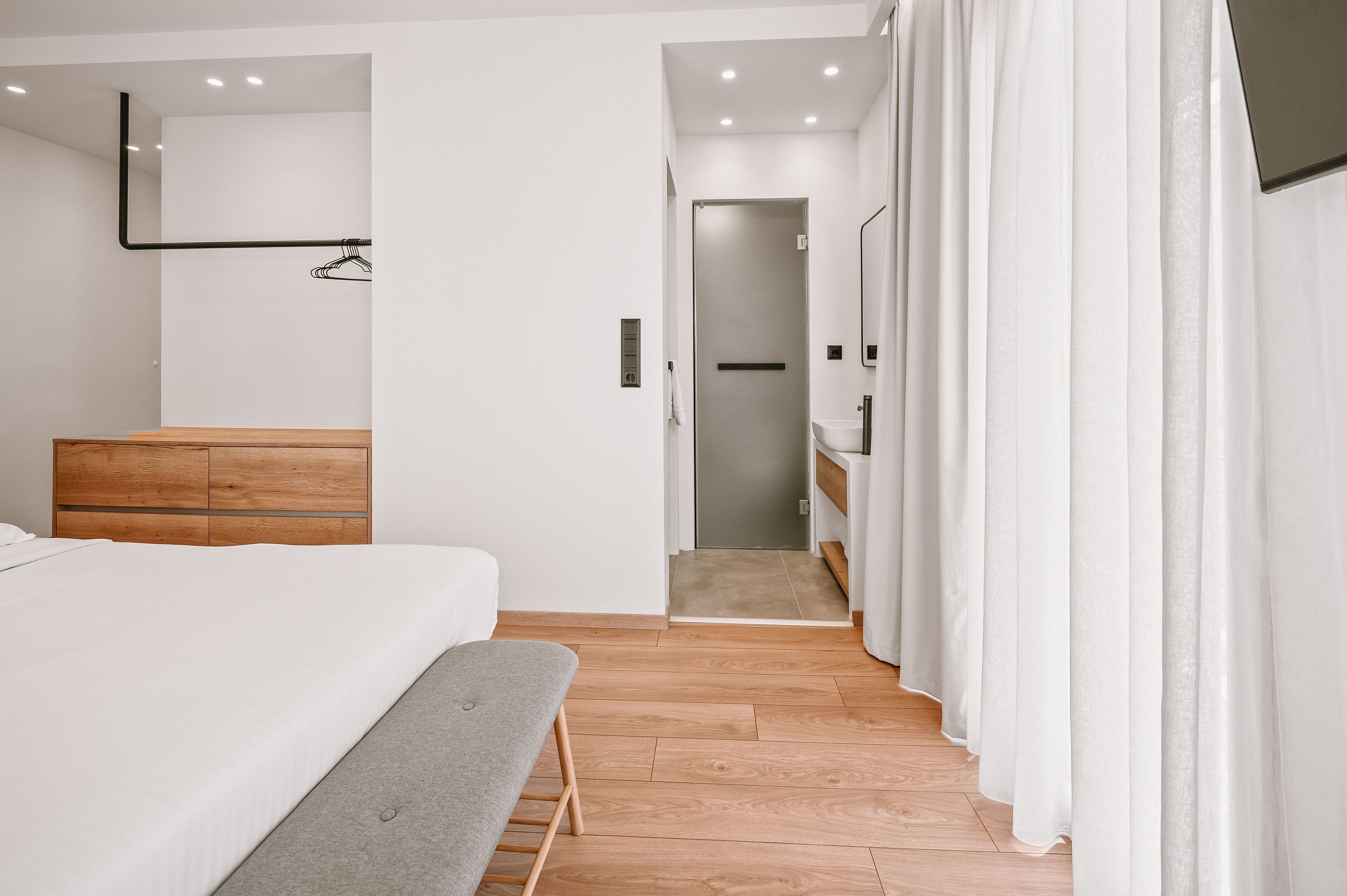 Une chambre moderne avec un lit en bois, un banc, une table de chevet, un miroir et une salle de bain visible à travers une porte ouverte.
