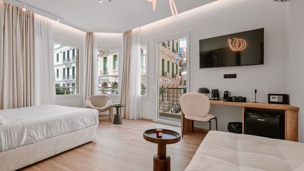 A modern hotel room with two beds, a desk, a TV, and a view of a building with balconies. - Plānis Str Urban Suites by Semavi (Heraklion)