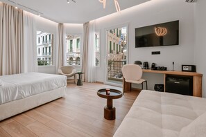 A modern hotel room with two beds, a desk, a TV, and a view of a building with balconies. - Plānis Str Urban Suites by Semavi (Heraklion)