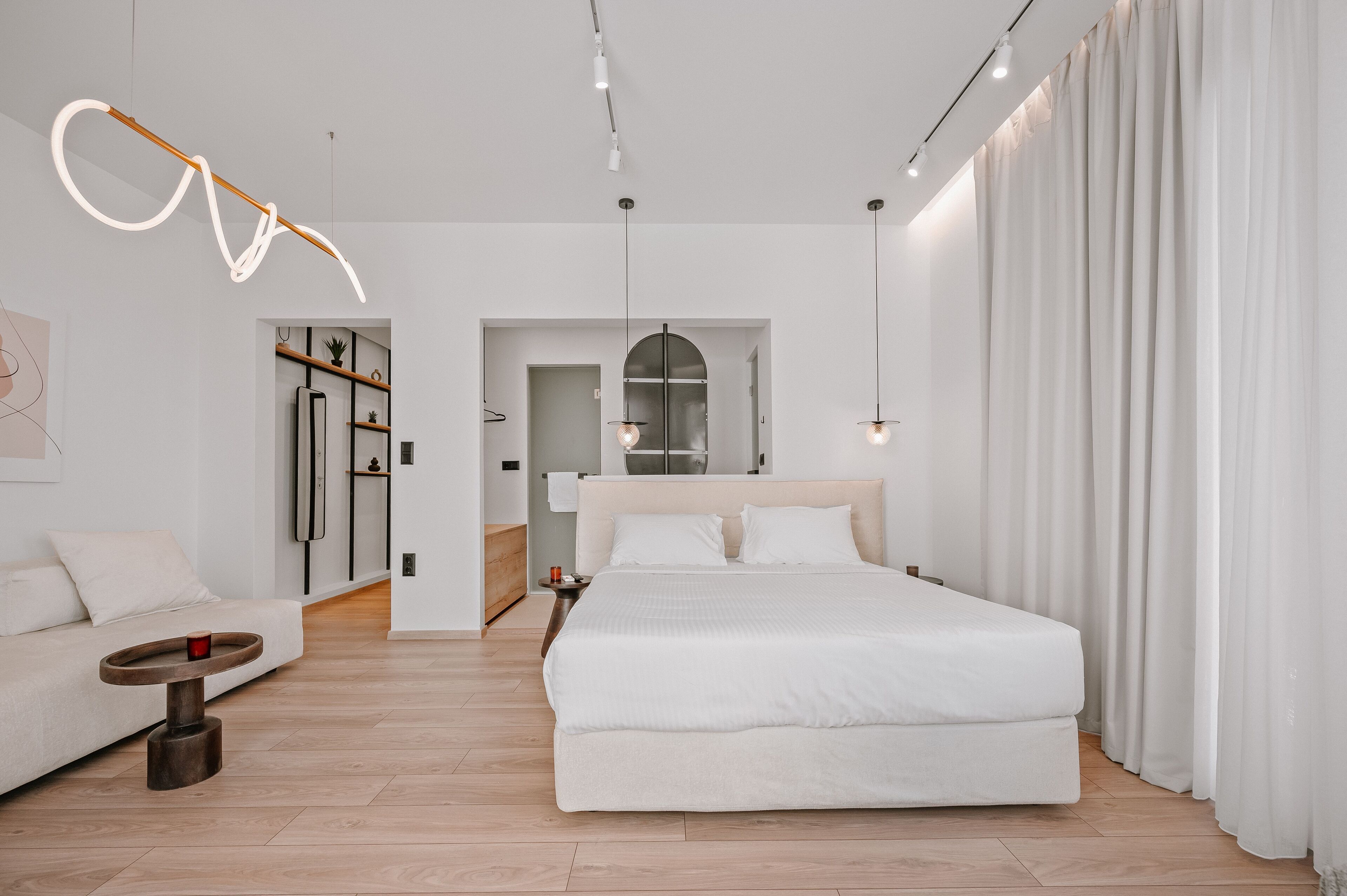 A modern bedroom with a large bed, a sofa, and a small table. There is a hallway leading to a bathroom and a kitchen area.