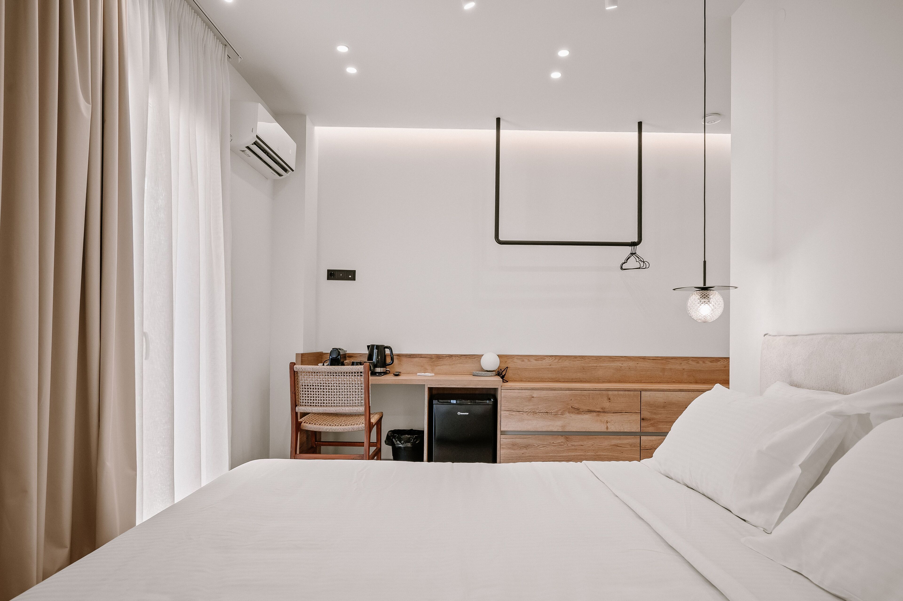 Une chambre d’hôtel moderne avec une tête de lit en bois, un bureau avec une chaise, un petit réfrigérateur et un lit recouvert de draps blancs.