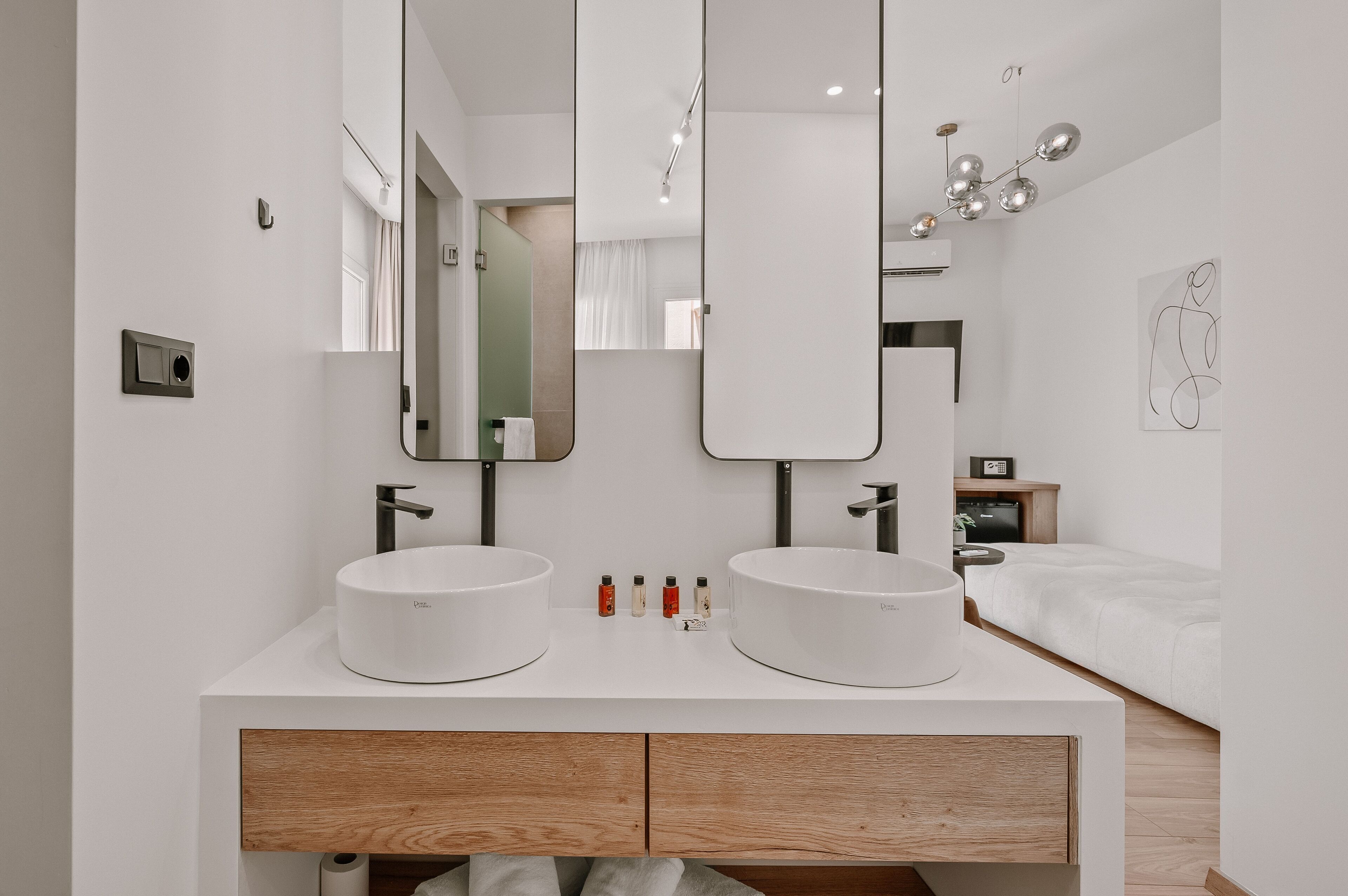 A modern bathroom with two sinks, a large mirror, and a bedside table with a lamp.