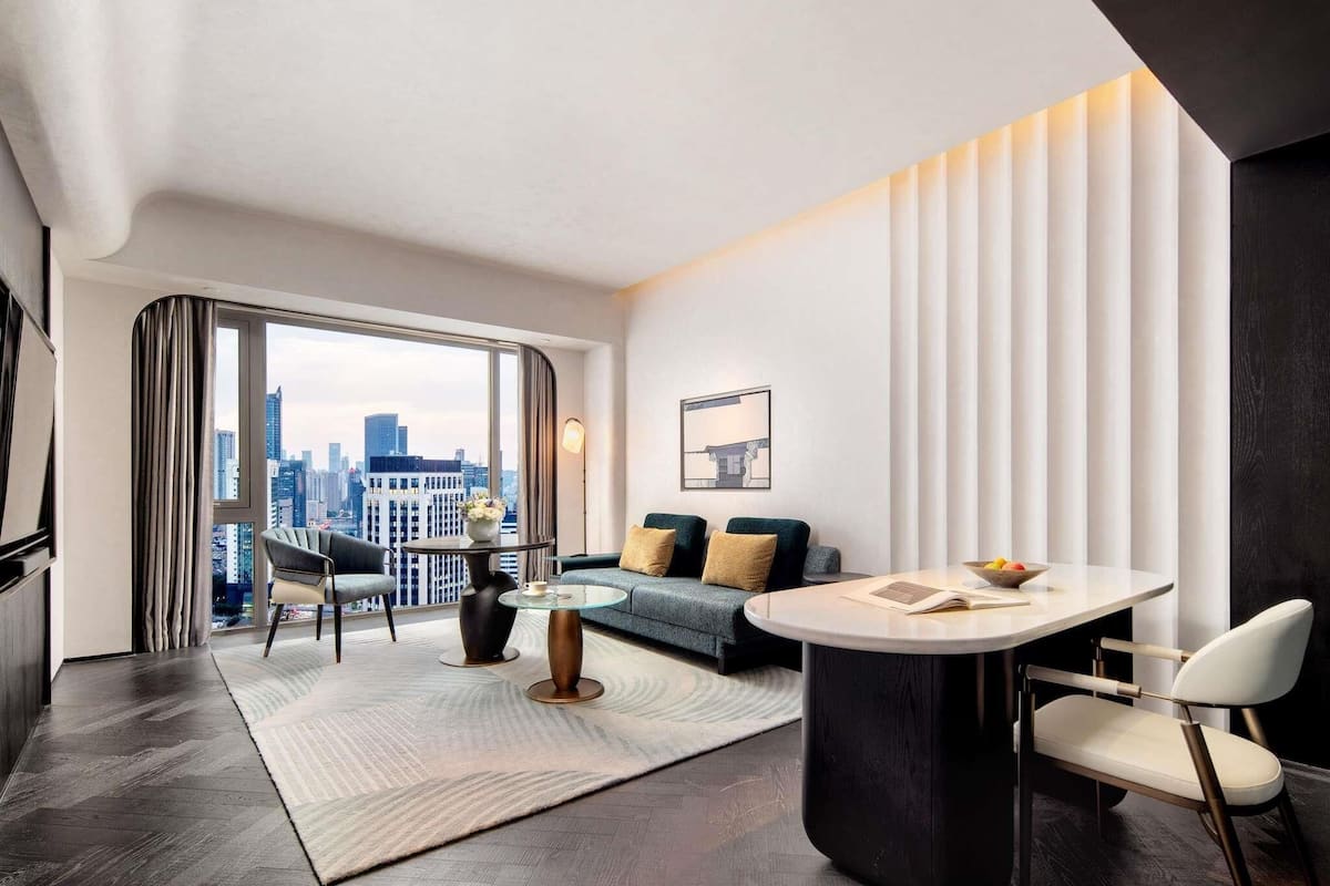 A modern hotel room with a large window, a round dining table, a sofa, and a chair.