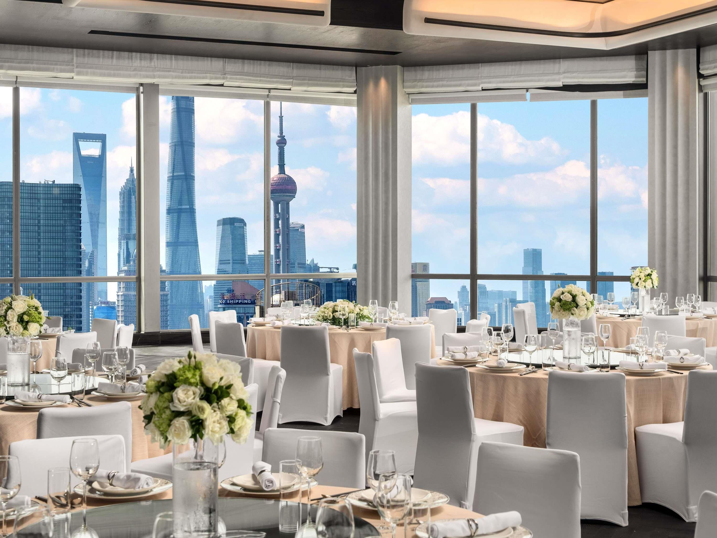 Un comedor de hotel con mesas dispuestas para una comida, amplias ventanas que ofrecen vistas del horizonte de la ciudad y un diseño de interiores moderno.