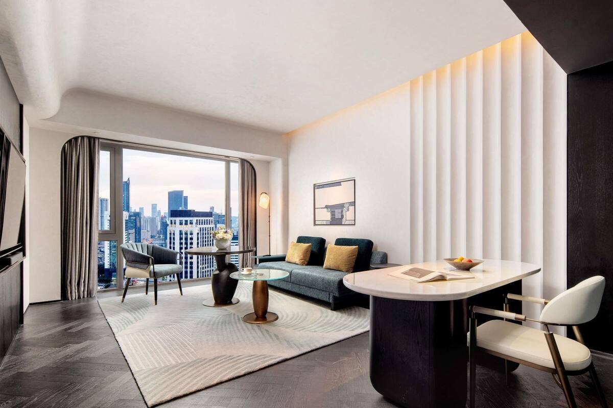 A modern hotel room with a large window, a round dining table, a sofa, and a city view.