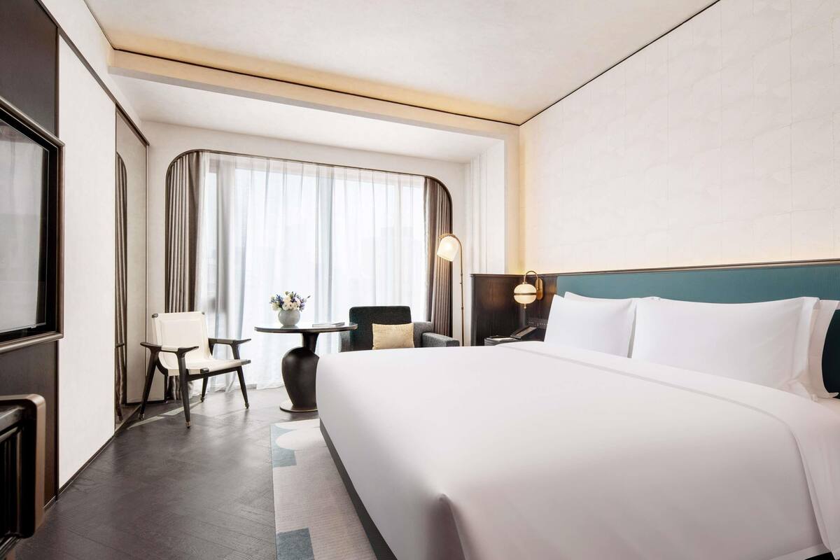 A hotel room with a large bed, a desk with a chair, a small table with a vase, and a large window with curtains.