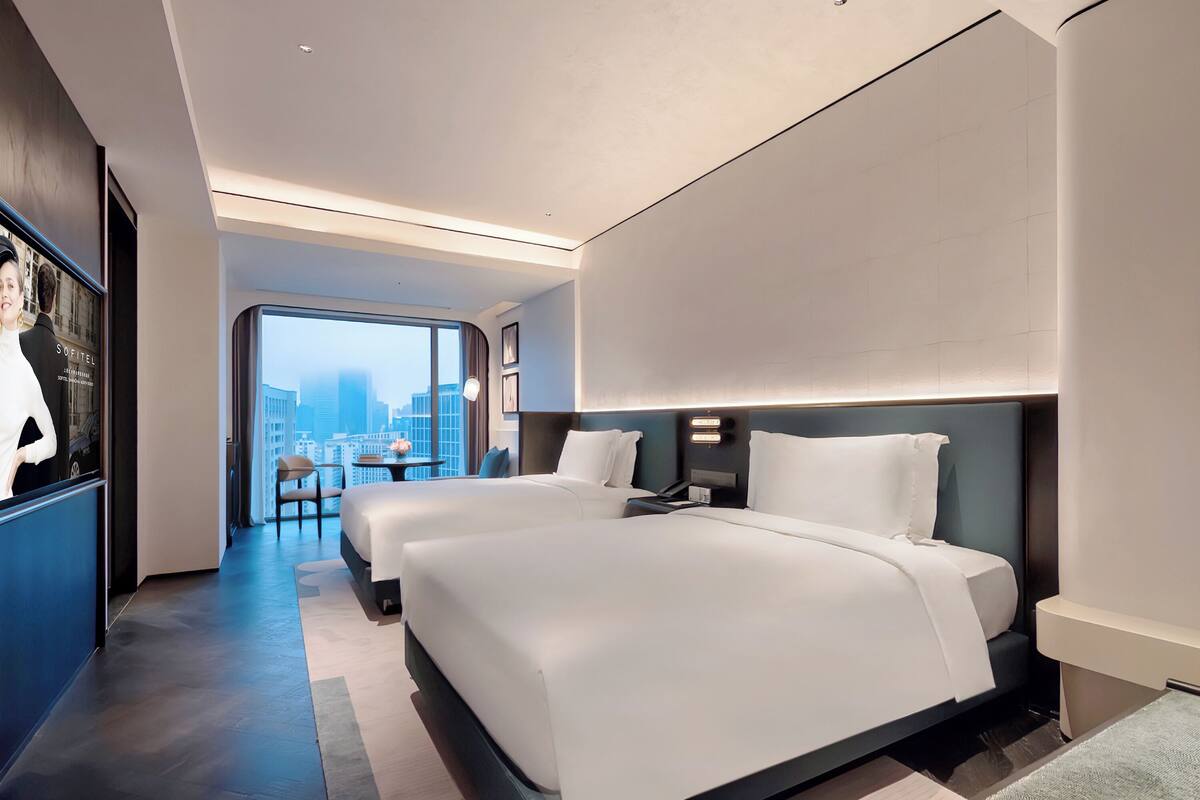 A modern hotel room with two beds, a desk, and a view of the city.