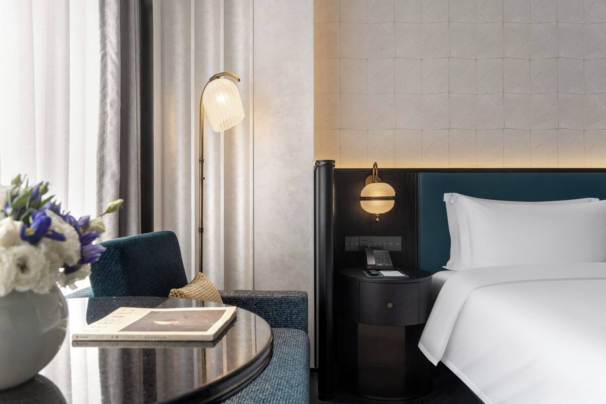 A hotel room with a bed, bedside table, lamp, chair, and a vase with flowers.