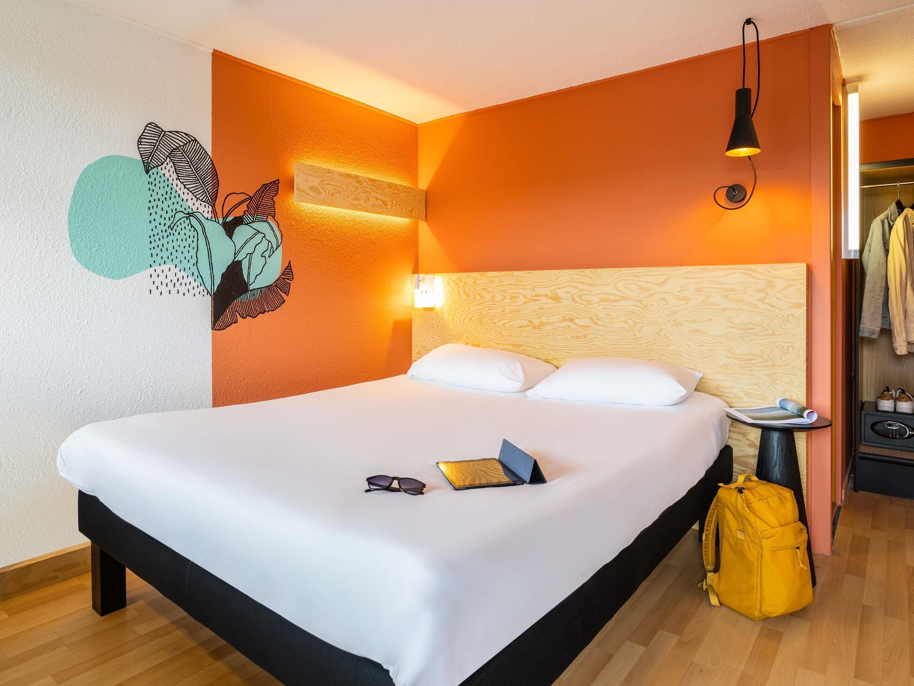 Et hotelværelse med en seng, et kunstværk på væggen, en sengetårnslampe, et lille bord med en bog og solbriller, samt en gul rygsæk.
