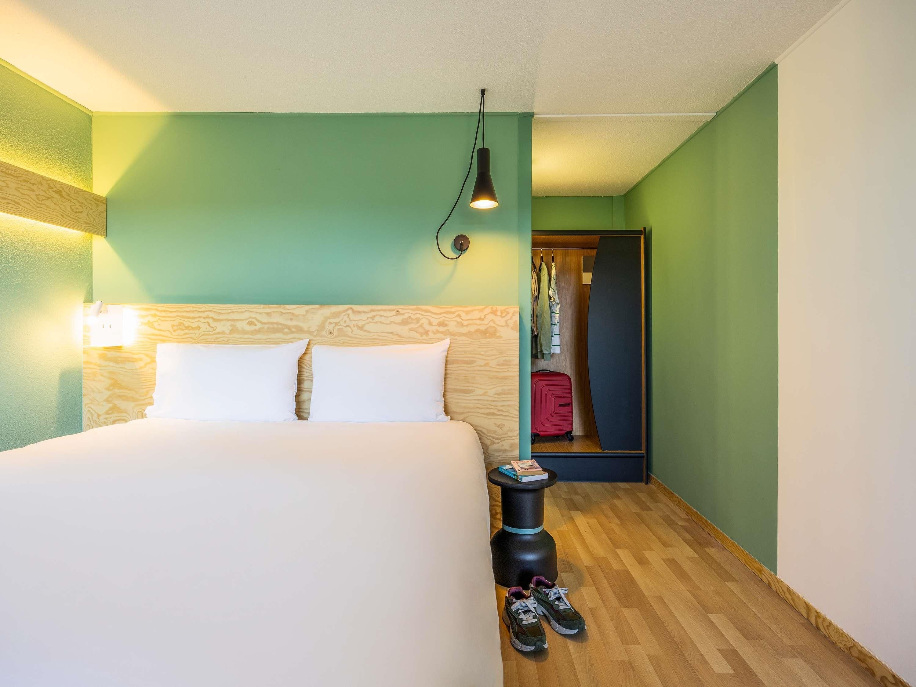 Une chambre d’hôtel avec un lit, une table de chevet, une lampe, une valise et une armoire où sont suspendus des vêtements.