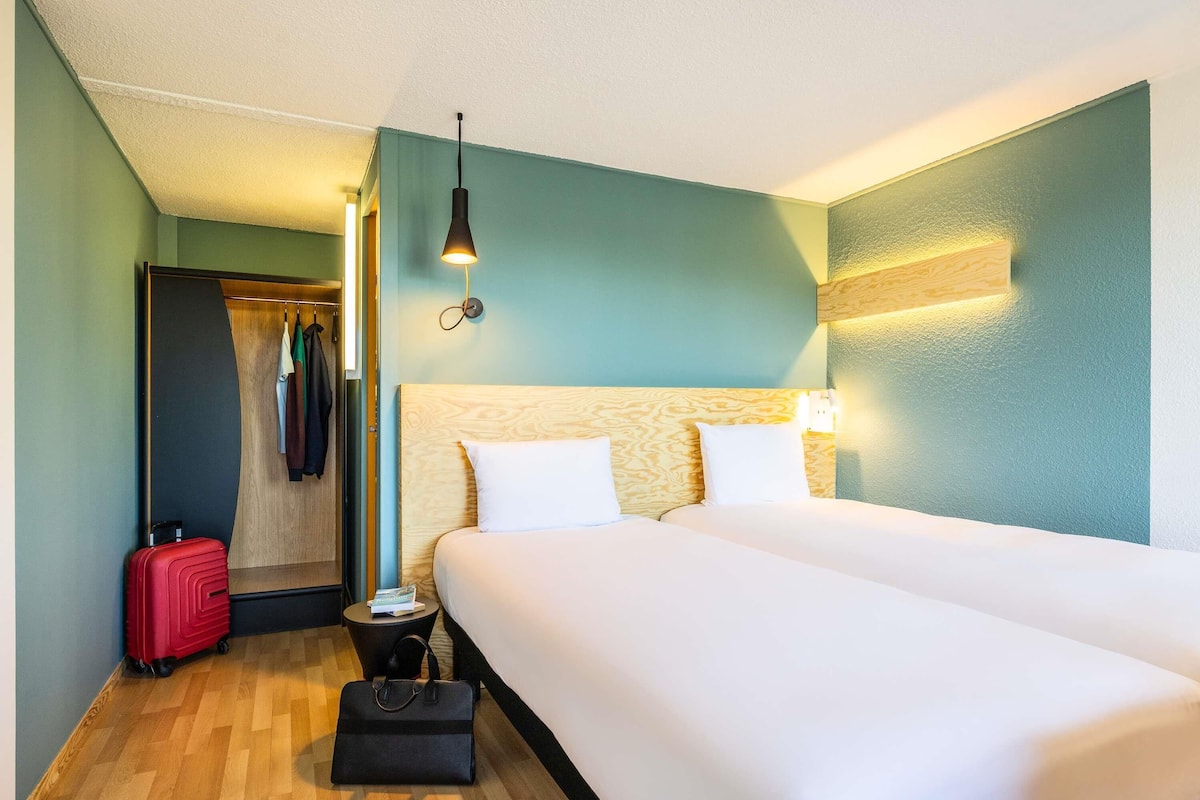 Una habitación de hotel con una cama grande, un armario, una maleta, un bolso de mano y una mesita de noche.