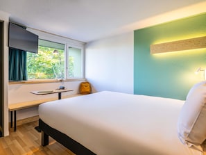 Habitación de hotel con cama, escritorio, silla, televisor, ventana con cortinas y lámpara.