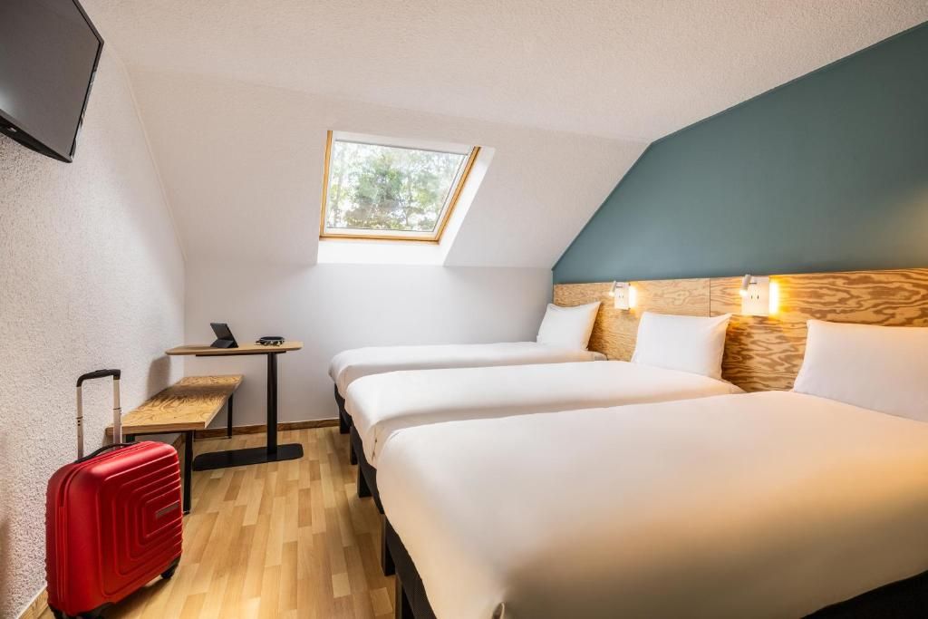 Een kamer met twee bedden, een klein tafeltje, een laptop, een rode koffer en een raam met uitzicht op bomen.