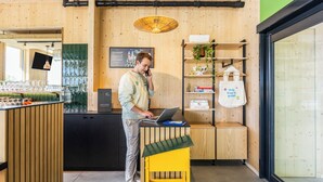 A person using a laptop at a wooden counter with a green-tiled backsplash, a hanging plant, and a white tote bag with text. - greet Brussels Airport Zaventem (Zaventem)