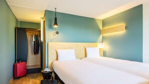 A hotel room with a double bed, a small bedside table, a red suitcase, and a closet with hanging clothes. - greet Brussels Airport Zaventem (Zaventem)