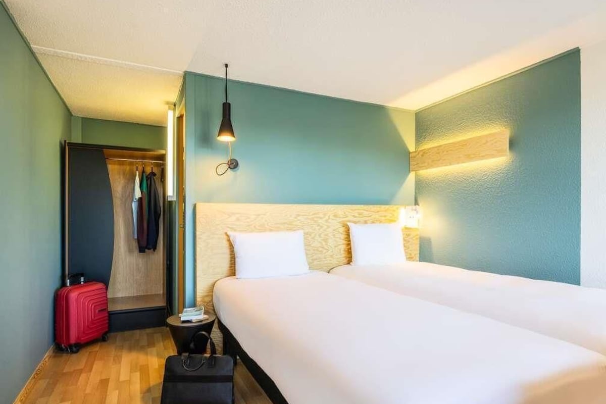 A hotel room with a double bed, a small bedside table, a red suitcase, and a closet with hanging clothes.