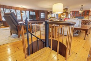 Uma escada de madeira com corrimão, uma sala de estar com sofá, televisão e uma área de jantar com mesa e cadeiras.