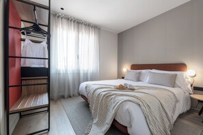 A neatly made bed with a white comforter and pillows, a bedside table with a lamp, a window with sheer curtains, and a clothing rack with a white top and a black top. - Arrivo Apartments (Barcelona)