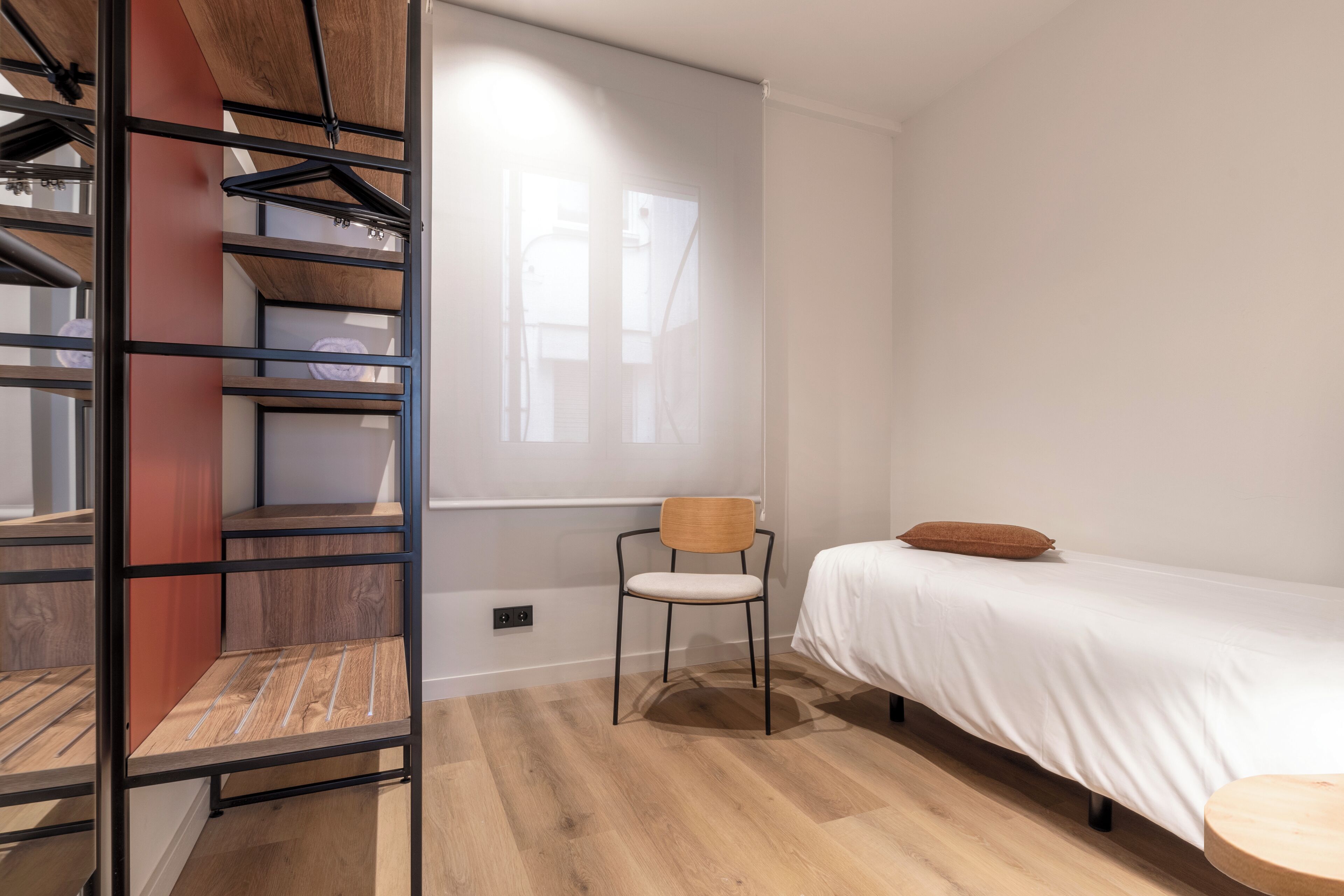 Un dormitorio con cama, una silla, piso de madera, armario y ventana.