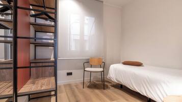 Un dormitorio con cama, una silla, piso de madera, armario y ventana.