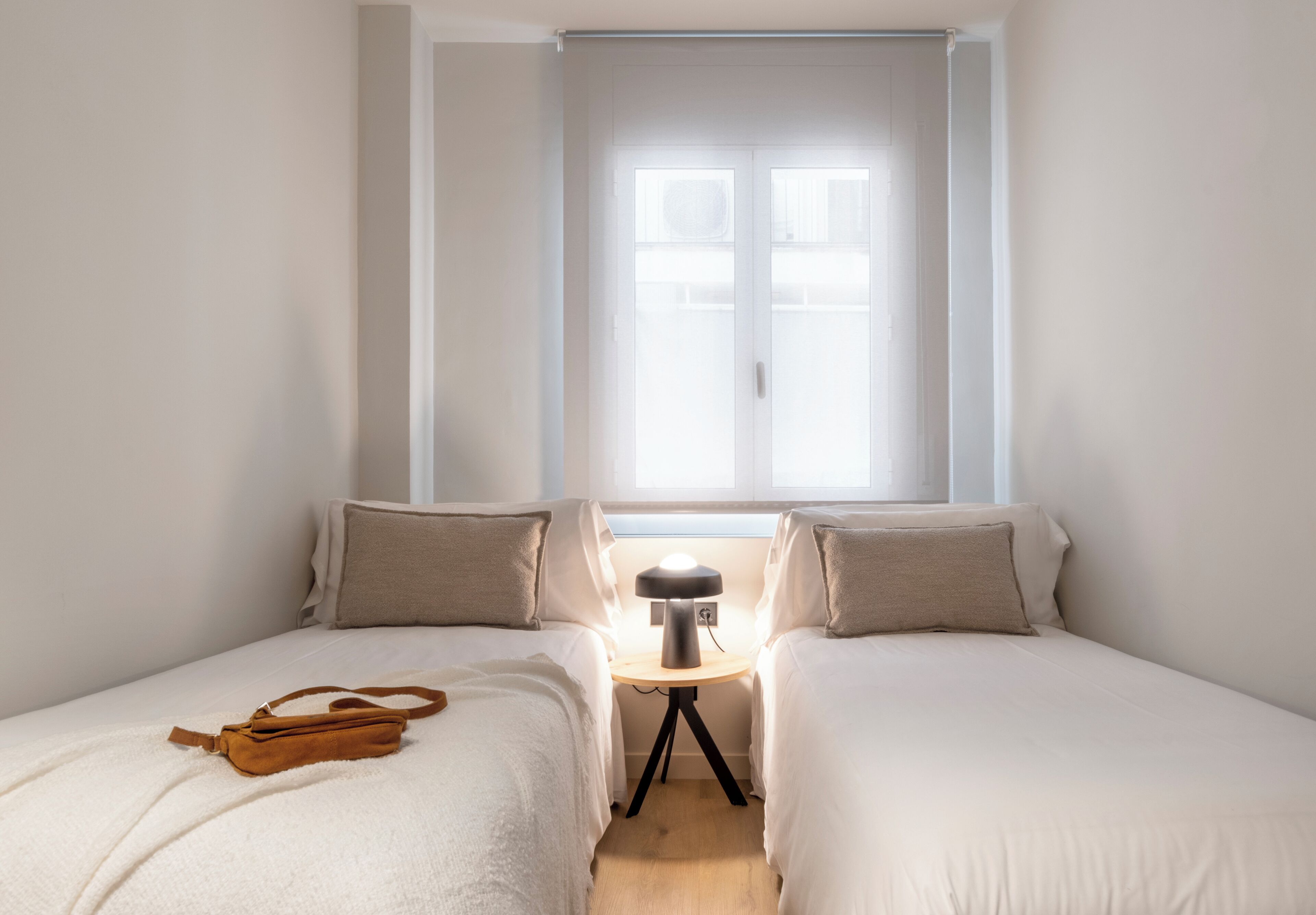 A bedroom with two beds, a bedside table with a lamp, and a window with blinds.