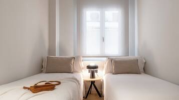 A bedroom with two beds, a bedside table with a lamp, and a window with blinds.