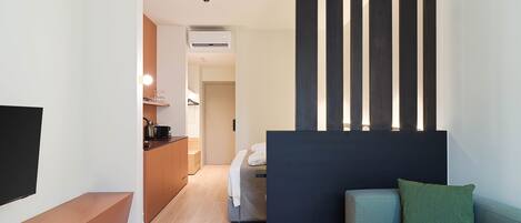 A modern hotel room with a flat-screen TV, a sofa with a green pillow, a wooden bench, a bed with a white blanket, a kitchen area, and a dark partition.