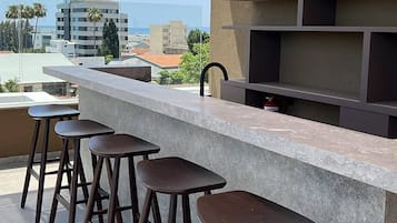 An outdoor bar area with a concrete countertop, wooden stools, hanging pendant lights, and a view of the cityscape.