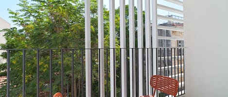 A balcony with two red chairs and a table, surrounded by white vertical blinds, overlooking a green area with trees and a building in the background.