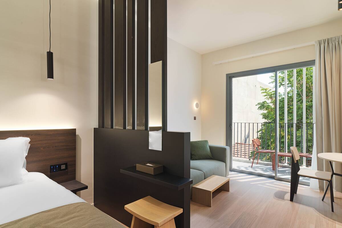 A modern hotel room with a bed, a desk, a chair, a small table, and a balcony with a view of trees.