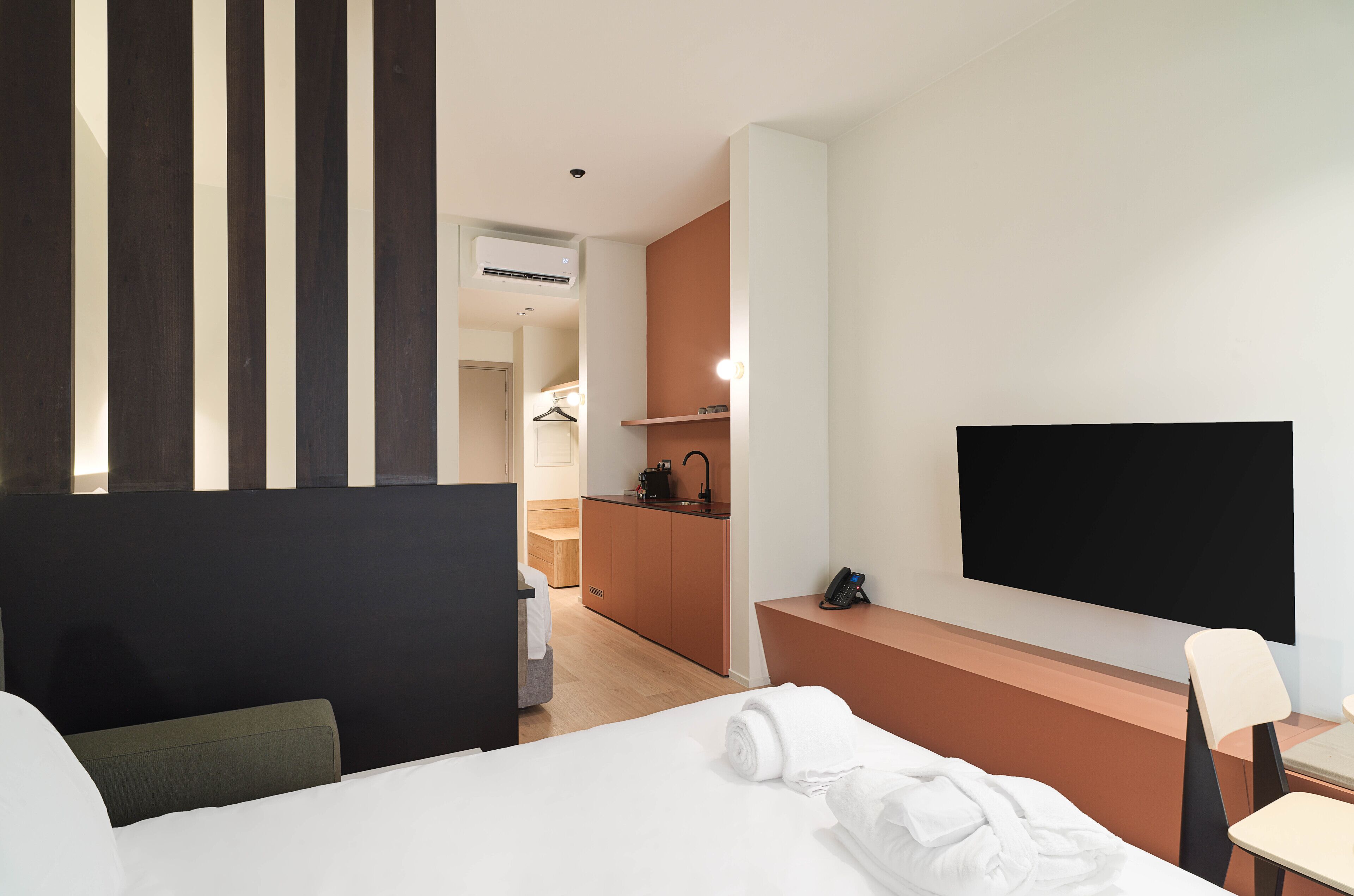 A hotel room with a bed, a desk, a chair, a TV, a kitchen area, and a bathroom.