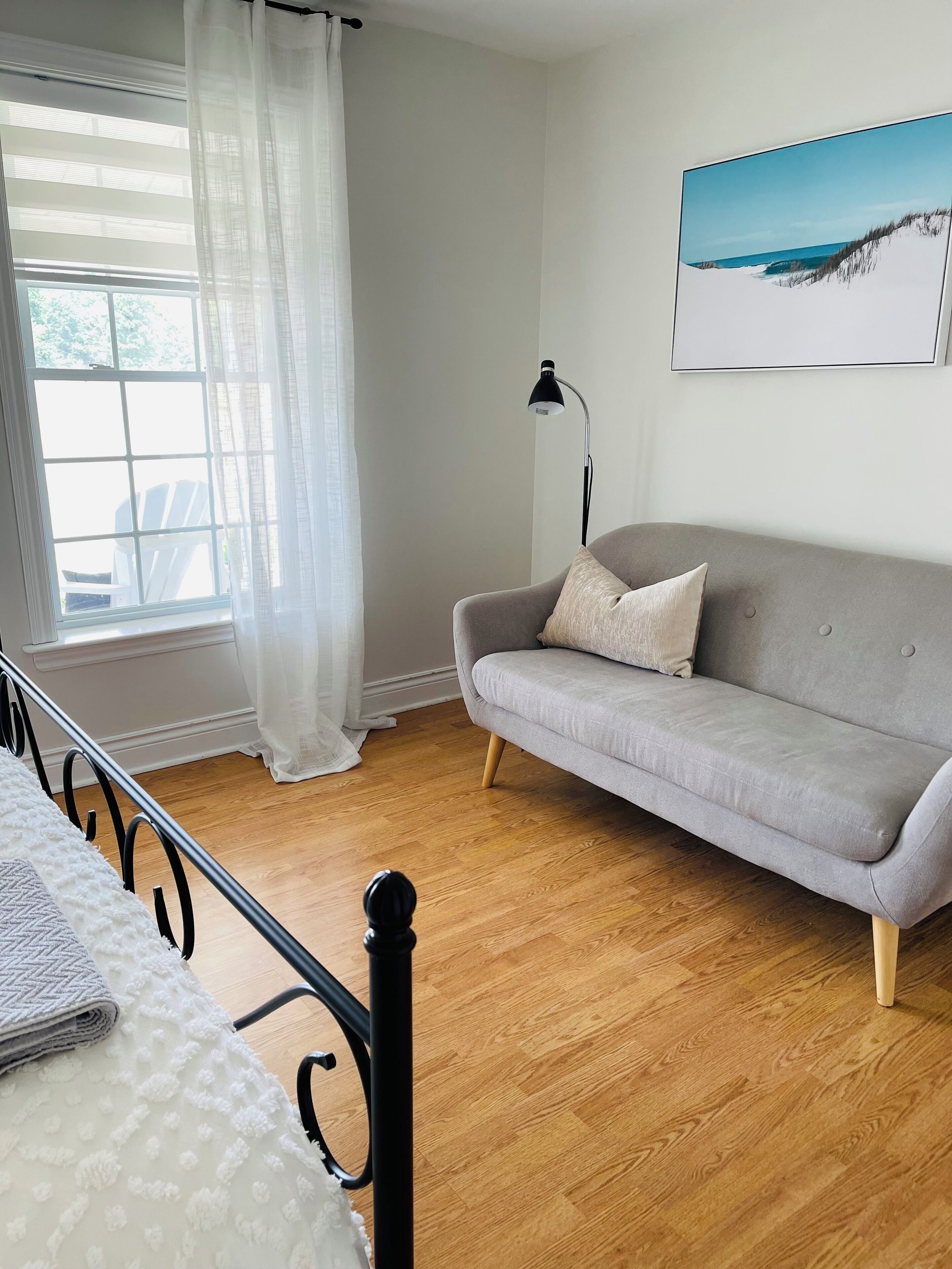 A room with a bed, a grey sofa, a floor lamp, and a painting of a beach scene.