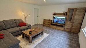 A modern living room with a grey sectional sofa, a wooden coffee table, a floor lamp, a TV on a wooden stand, and a wardrobe. - Haus am Wald Ruhig Gelegene Ferienwohnung mit Großem Garten (Bad Tabarz)