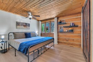 Ein Schlafzimmer mit einem Bett, einem Deckenventilator, einem Nachttisch, einem Fenster mit Vorhängen und einem Bücherregal.