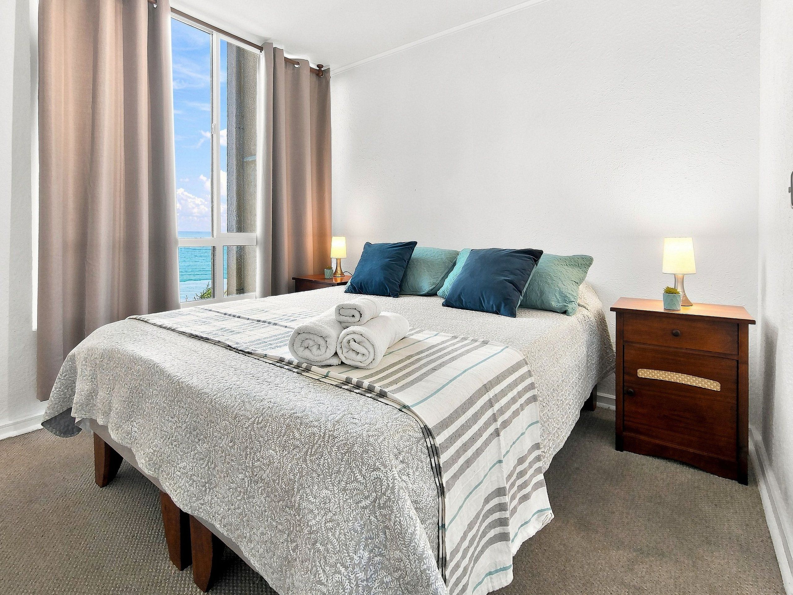 Habitación de hotel con cama, dos mesitas de noche, una ventana con cortinas y vista al mar.