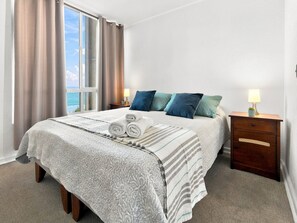 A hotel room with a bed, two bedside tables, a window with curtains, and a view of the sea.