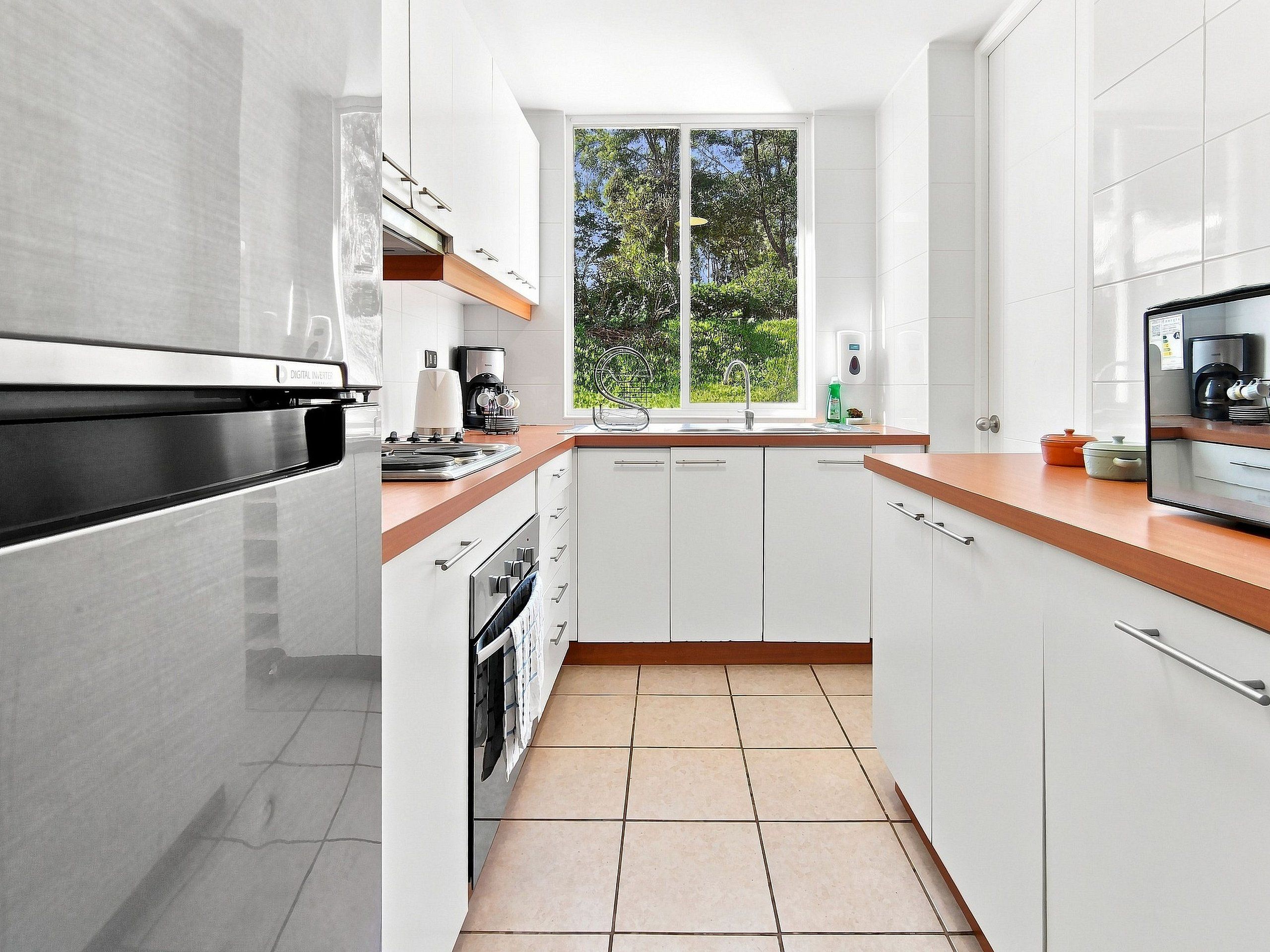 A modern kitchen with white cabinetry, a stainless steel refrigerator, a built-in oven, and a window providing a view of greenery outside.