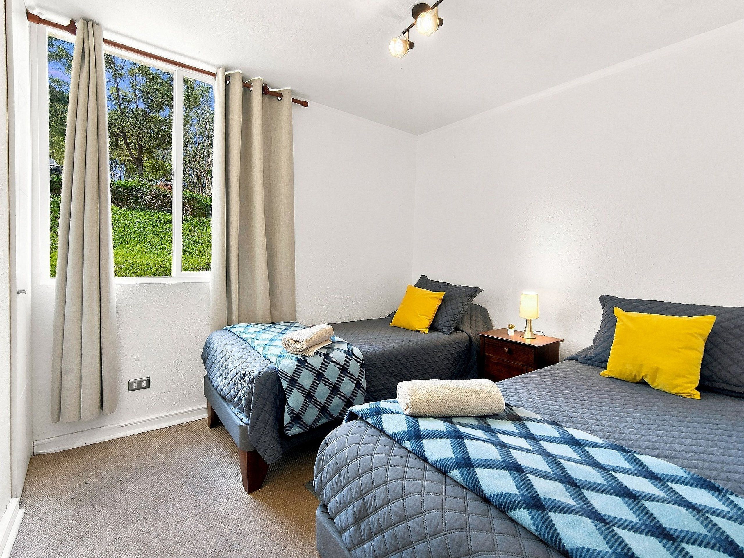 A room with two beds, a nightstand with a lamp, and a window with curtains.