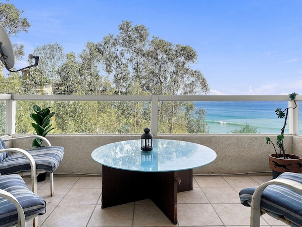 A round glass table with a metal base, surrounded by two striped chairs, on a balcony with a view of the ocean and trees.
