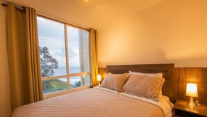 A hotel room with a large bed, a nightstand with a lamp, and a window with curtains. - Oceana Suites Costa Quilen (Puchuncavi)