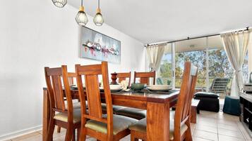 Uma sala de jantar com mesa e cadeiras de madeira, um quadro na parede e vista através de grandes janelas.