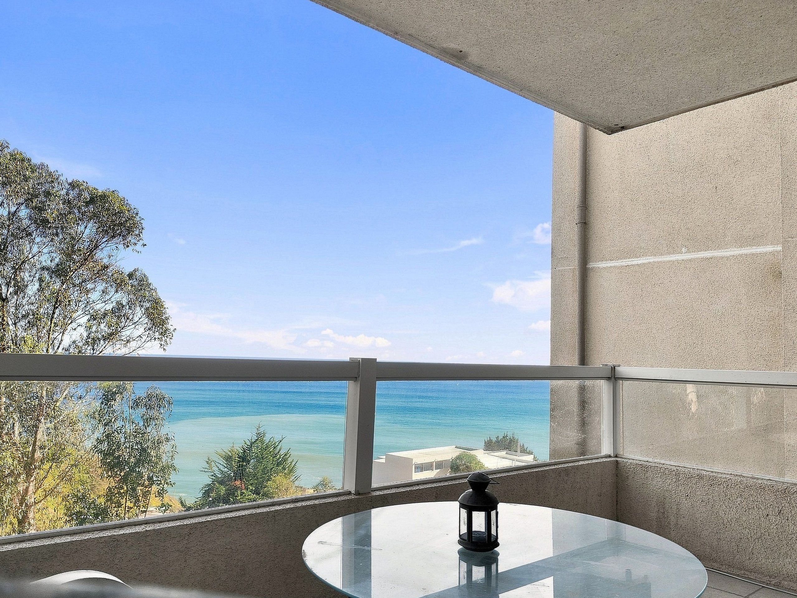 A balcony with a glass table and a lantern, overlooking a clear blue sea and distant buildings.